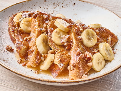 Thick-cut, custard-dipped challah bread griddled and topped with fresh sliced bananas, pecans and caramel sauce and lightly dusted with powdered cinnamon sugar. 