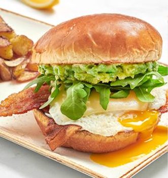 Bacon, egg, cheese and avocado on a brioche bun on a plate with seasoned potatoes.